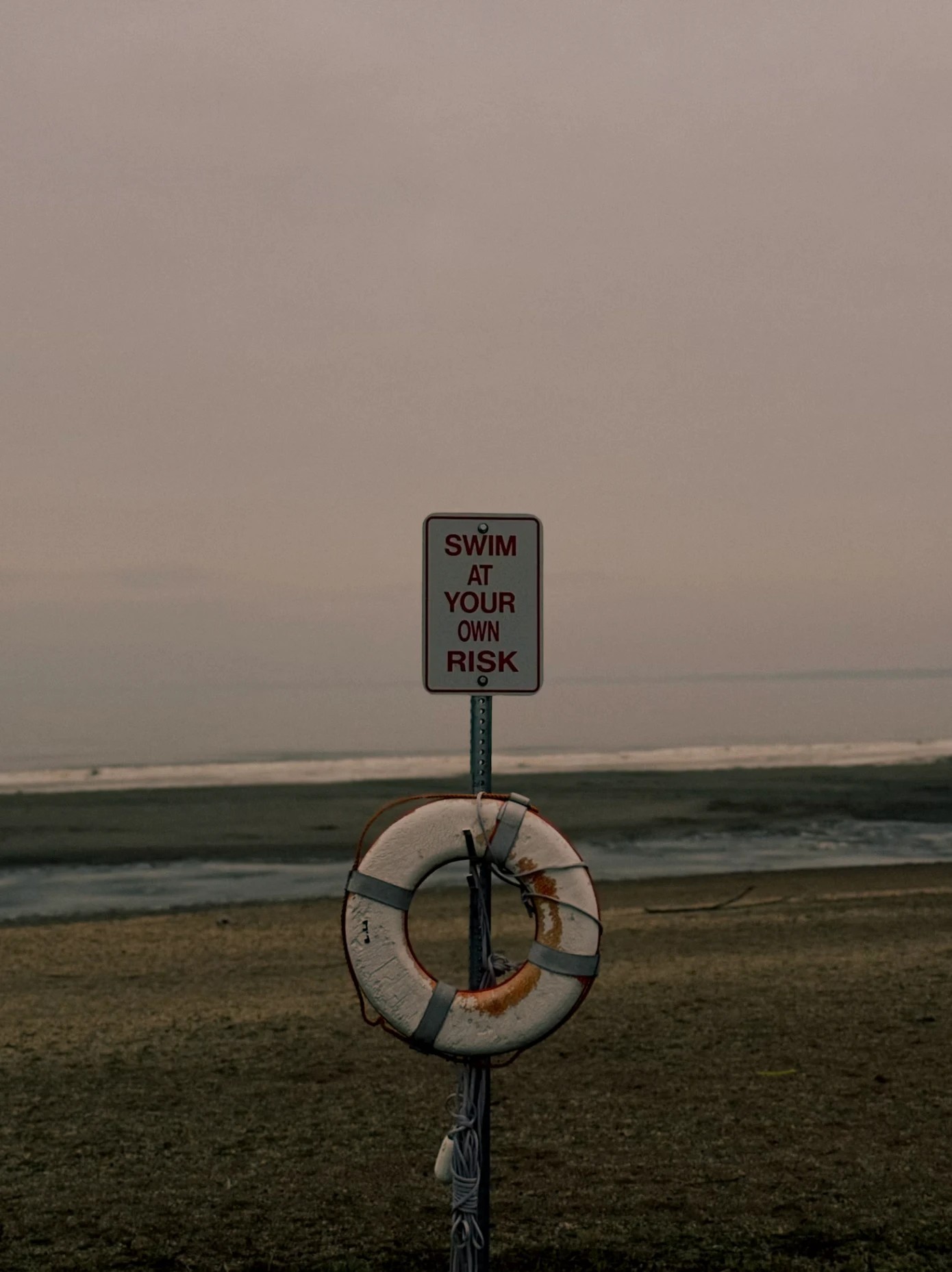 A lifebuoy and 'Swim at Your Own Risk' sign on an empty beach